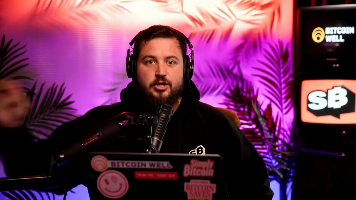 The image features a man wearing headphones, sitting at a desk with a laptop computer open in front of him. He appears to be speaking into a microphone or recording device, possibly participating in an interview or podcast discussion about Bitcoin. The background includes a TV screen displaying "SBS" and "bitcoin wells", suggesting that the conversation is taking place on a television set.