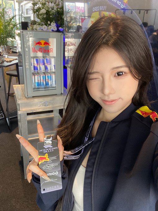 A young woman with long dark hair winks playfully at the viewer while holding up a Red Bull-branded event credential, dressed in a **Red Bull Racing** jacket. Behind her, a Red Bull branded mini-fridge displays the words "Red Bull," and the credential reads **"PADDOCK CLUB"** with a Singapore Grand Prix 2024 design. The setup reflects her day enjoying F1 festivities in Singapore alongside a JJ Lin concert.