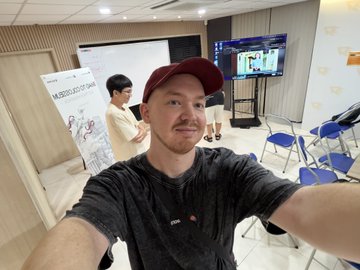 A man wearing a red cap takes a selfie in a brightly lit room, with other people and a display monitor visible in the background.