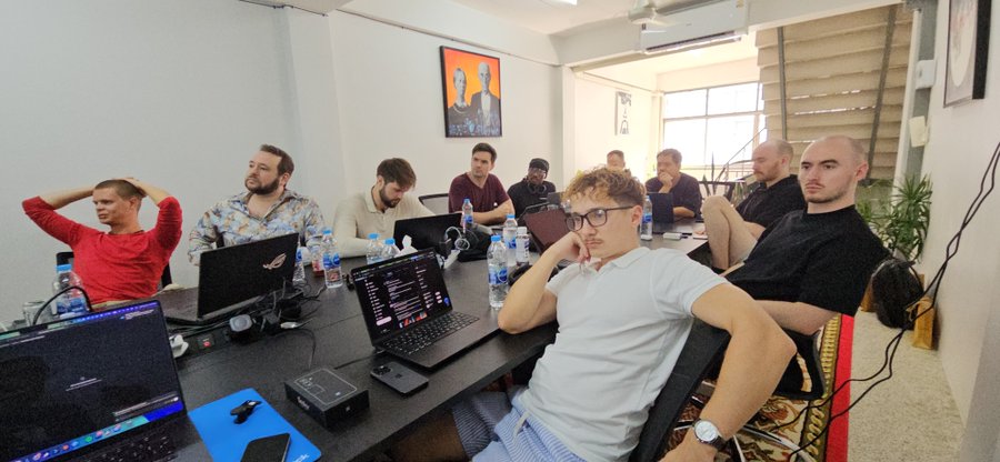 A group of men sit around a conference table in a brightly lit room, working on laptops with one man in a white polo shirt and glasses looking directly at the camera.