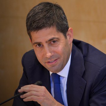 A middle-aged man in a dark suit and blue tie speaks into a microphone, photographed in what appears to be a formal hearing or press setting.
