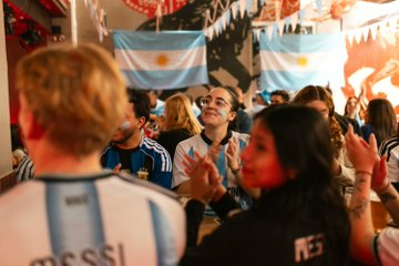 A crowd of fans, many wearing Argentina jerseys with "MESSI" visible on one, celebrate under hanging Argentine flags and festive decorations.