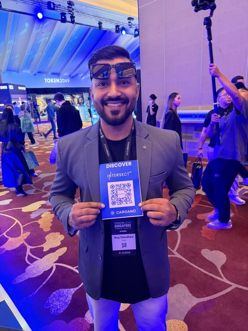 A man in a grey blazer and sunglasses smiles at the TOKEN2049 Singapore event, holding a Cardano 'DISCOVER INTERSECT' QR code card.