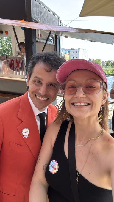 A selfie of a smiling man in a red suit with a 'RAVE' badge and a woman wearing a pink cap and sunglasses, with water visible in the background.