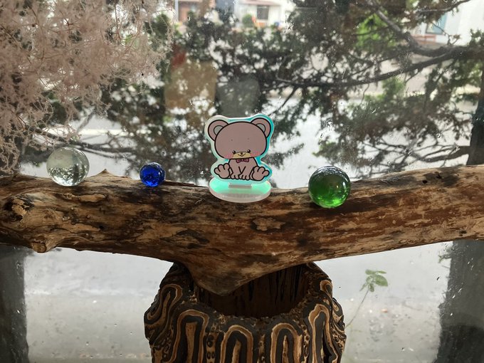 A small cartoon bear figurine sits on a wooden branch/log, accompanied by three glass marbles (clear, blue, and green), with trees visible through a window in the background.
