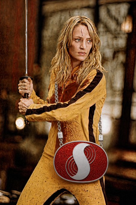 A blonde woman in a yellow costume holds a sword while standing with chains and a red circular emblem; a red-and-white mark is visible on the emblem.