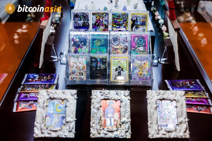 A display of various collectible trading cards and figures on a dark table with a 'bitcoin asia' watermark in the corner.