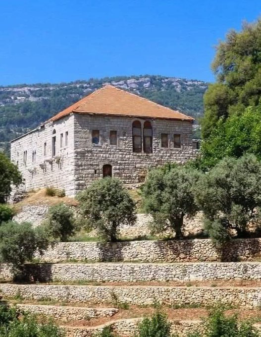 A large, two-story stone building with an orange tiled roof sits on a hillside, surrounded by terraced stone walls, olive trees, and green vegetation under a clear blue sky.