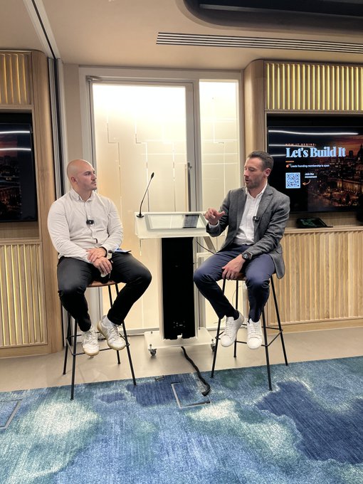 Two men in business casual attire sit on stools in a studio setting, appearing to engage in an interview or discussion, with a screen behind them displaying "Let's Build It".