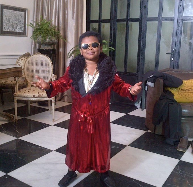 A person of short stature wearing a red velvet robe with black fur collar and dark sunglasses, standing with arms outstretched on a black and white checkered marble floor in an elegant room with classical furniture.