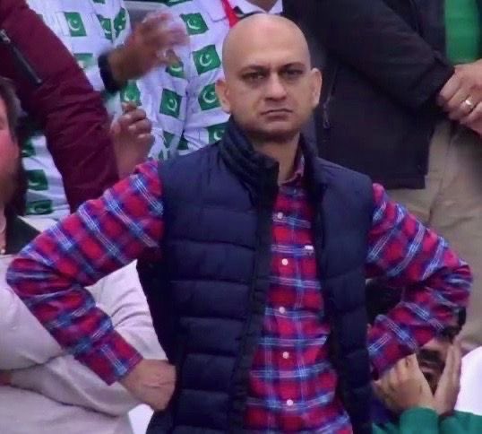 Bald man wearing a red and blue plaid shirt under a navy puffer vest, standing with hands on hips at what appears to be a sporting event with people in green and white attire in the background.
