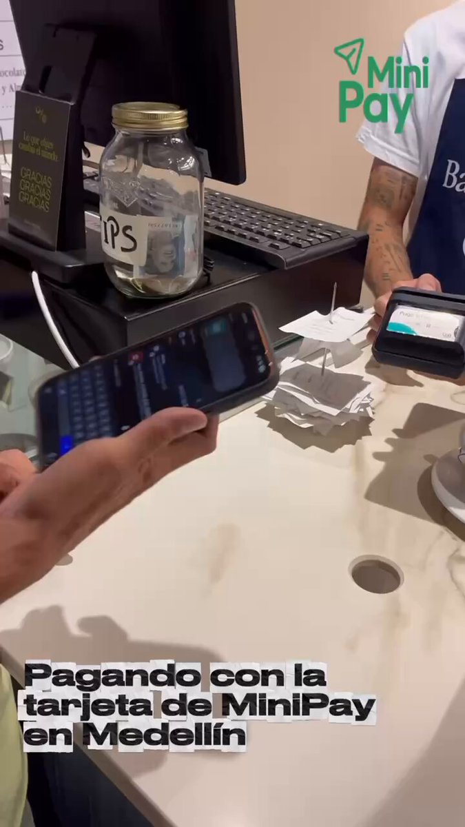 The image shows a person using their phone at a payment terminal, likely a MiniPay device, while another person is standing behind them. The scene takes place in a coffee shop setting with a table visible in the background. A bottle can also be seen on the table. The main focus of the image is the interaction between the two individuals and the use of technology for making payments at the terminal.