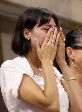 A young woman with shoulder-length dark hair covers her face with both hands, displaying a diamond ring on her finger and a gold bracelet on her wrist. Her emotional reaction suggests an engagement, tying into the celebratory post announcing the exciting news with tags to friends.