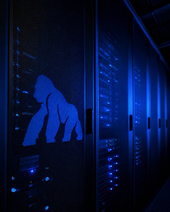 A row of illuminated server racks bathed in blue light displays the iconic APE (ApeCoin) logo on the front mesh panel, symbolizing the cryptocurrency's digital infrastructure. The post's themes of "Access, Reliability, Security" align with the image's depiction of secure, operational server hardware—reinforcing $APE's presence in the blockchain ecosystem.