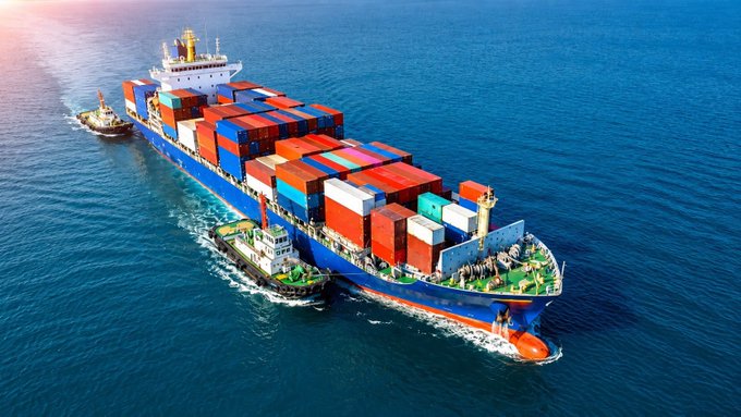 An aerial view shows a large cargo ship loaded with shipping containers being escorted by smaller tugboats across the ocean.