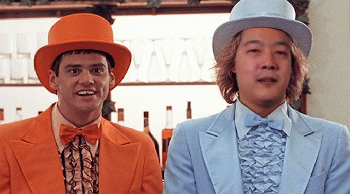 Two men stand side by side in the image, one wearing a vibrant orange suit with a matching top hat and bow tie, and the other dressed in a light blue suit with a coordinating top hat and bow tie, with "HALVA" partially visible on a sign behind them. The caption "That confirmation time, it's fabulous!" humorously nods to the absurdity of waiting for a transaction to be confirmed on the blockchain, comparing the experience to the over-the-top, stylish attire of the duo from the iconic 1997 film *Dumb and Dumber*.