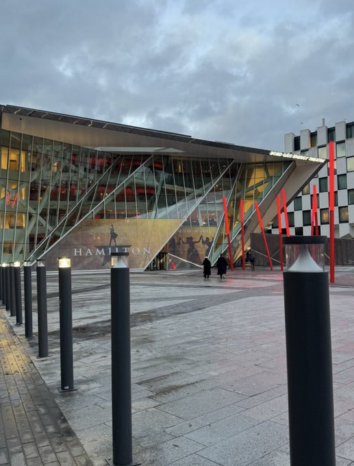 An urban plaza at dusk with black bollards in the foreground and a modern glass building in the background displaying a 'HAMILTON' musical advertisement.