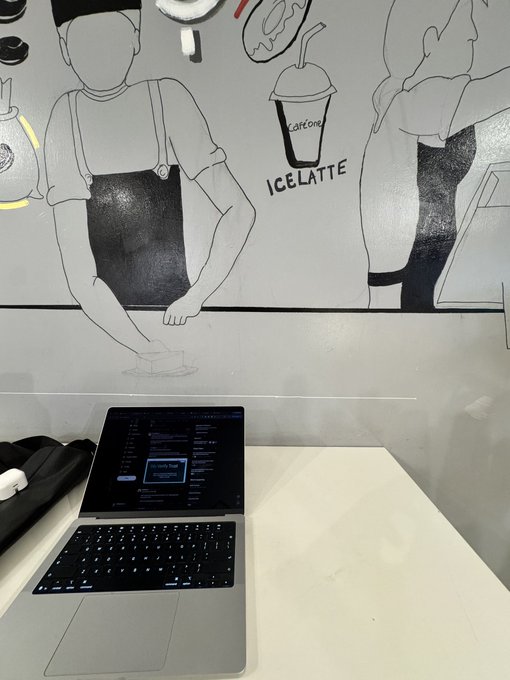 A MacBook laptop open on a white counter displaying what appears to be a coding or chat interface, with a black-and-white wall mural of a barista and an "ICE LATTE" illustration in the background.