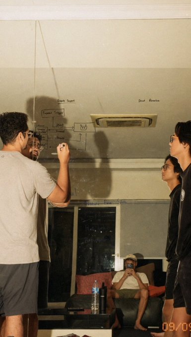 Several young men gathered around a whiteboard in a dimly lit room, with one drawing a diagram labeled with terms like "Picard's Theorem" and "Descartes," while another sits on a couch checking his phone.