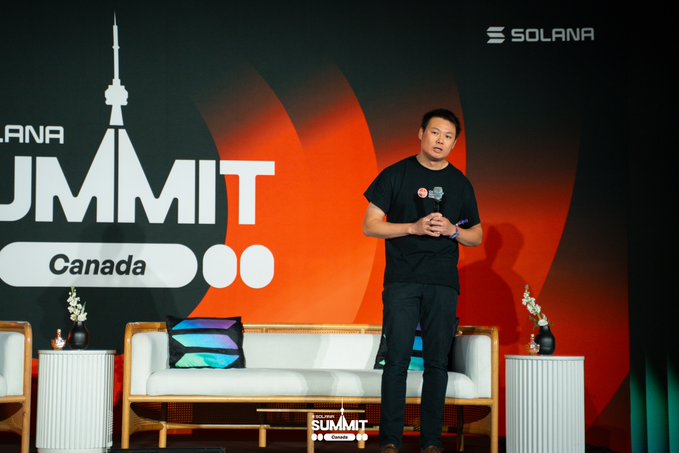 A man in a black t-shirt and dark pants stands on stage at the Solana Summit in Canada, speaking to an audience with a microphone in hand.