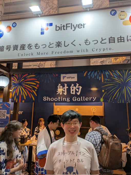 A man in a "Stablecoin Chat Calder" Japanese t-shirt smiles at the camera in front of the bitFlyer booth at what appears to be a crypto/Web3 conference, with signage reading "Unlock More Freedom with Crypto" alongside a "Shooting Gallery" promotional display decorated with fireworks imagery.