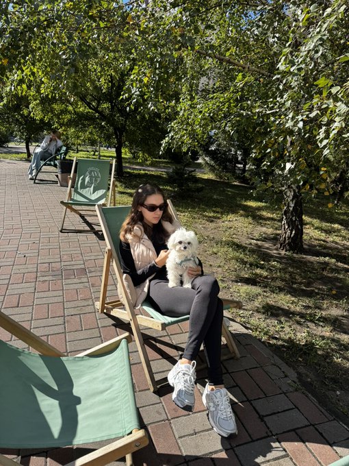 A woman wearing sunglasses sitting in a green deck chair outdoors holding a small white dog, with another person visible in the background.