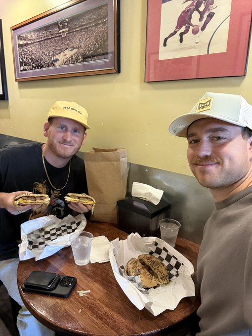 Two men sit at a wooden table eating sandwiches; one wears a yellow hat and shows his sandwich to the camera while framed sports photos hang on the wall behind them.