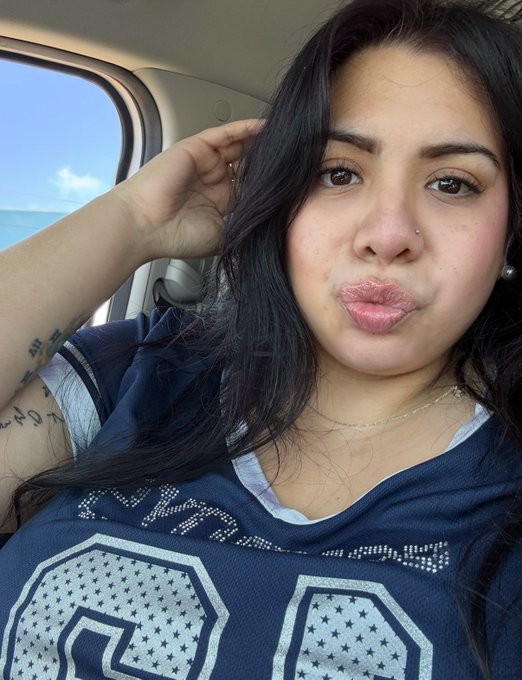 A close-up selfie of a woman with dark hair blowing a kiss toward the camera while sitting inside a vehicle, with the ocean visible through the window behind her.