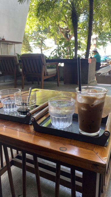 A wooden table at an outdoor café holding two black trays with iced coffee and glasses of water, with wooden chairs and greenery visible in the background.
