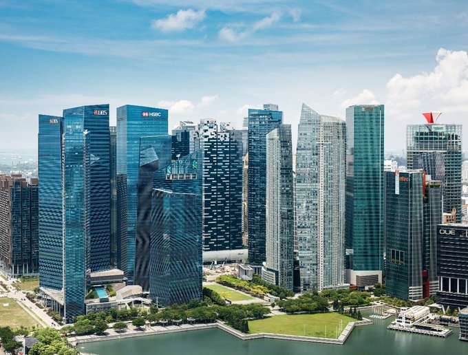 A skyline view of Singapore's Marina Bay financial district featuring skyscrapers like HSBC, OUE, and other corporate towers surrounding green park space and water.