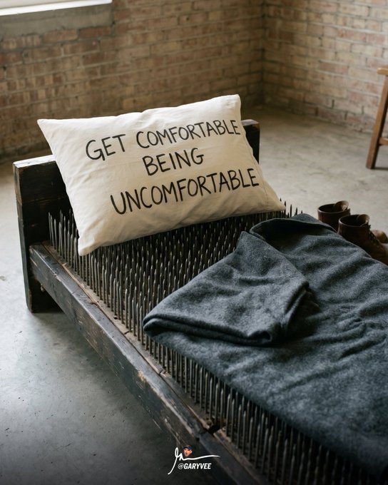 A DIY bed frame made of wood and nails features a pillow reading 'GET COMFORTABLE BEING UNCOMFORTABLE', covered with a grey blanket.