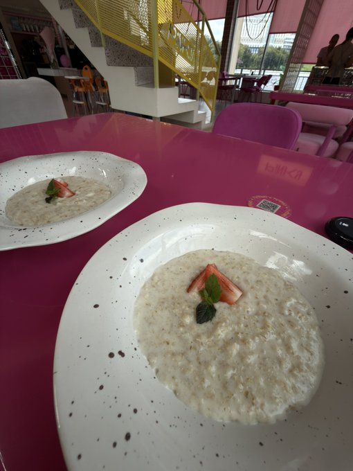 Two white plates of oatmeal or porridge topped with strawberry slices and mint leaves, served on a glossy pink table in a colorful restaurant setting.