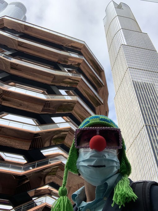 A person wearing a green knitted hat with tassels, a red clown nose, and a face mask poses for a selfie in front of The Vessel (the honeycomb-shaped copper structure) and 30 Hudson Yards skyscraper in New York City, perfectly capturing the playful, community-driven spirit of the "GMFriendly Friday" crypto shoutout celebration.