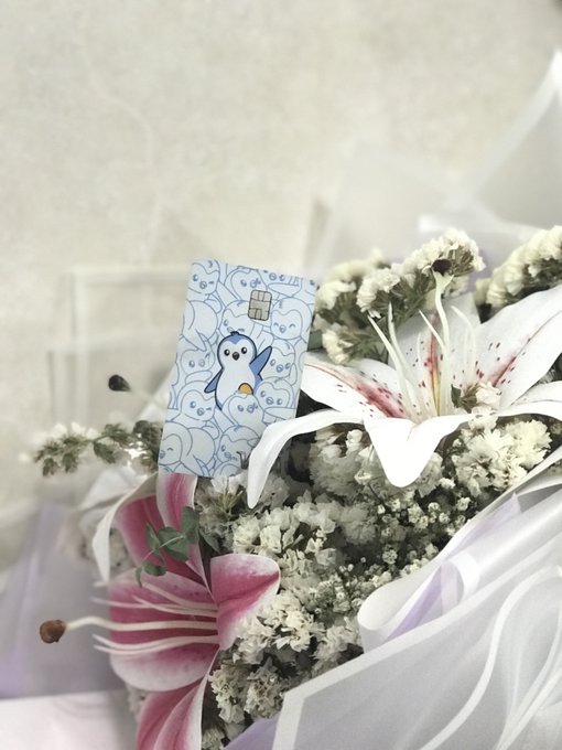 A close-up of a floral bouquet featuring white and pink lilies, baby's breath, and a decorative blue and white paper pick with a cute penguin illustration tucked among the flowers.