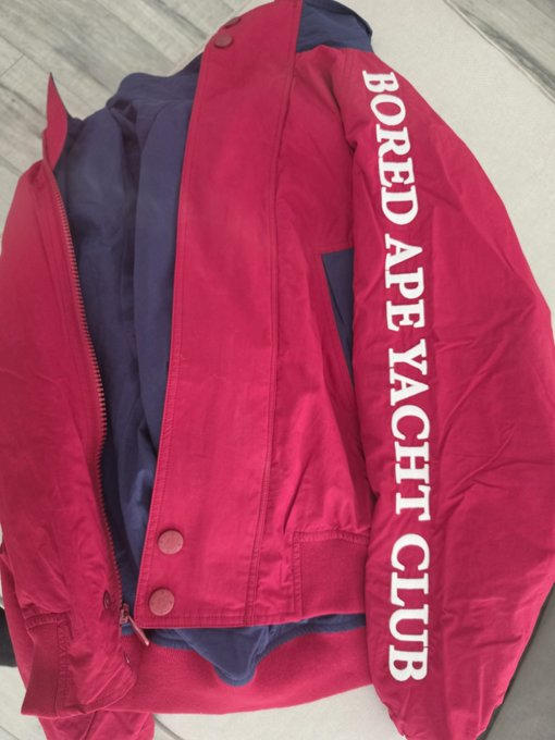 A red Bored Ape Yacht Club jacket laid flat, featuring the brand name in large white vertical text down the left sleeve and a navy blue interior lining.