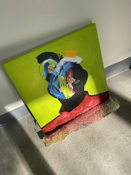 A brightly colored abstract painting on canvas resting on a paint-splattered floor, featuring bold green, blue, red, and yellow brushstrokes depicting a stylized figure.