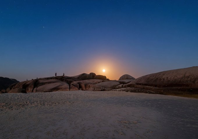 A nighttime landscape photo captures a bright moon rising over a rocky terrain, with a few small silhouetted figures standing on the rocks under a starry sky.