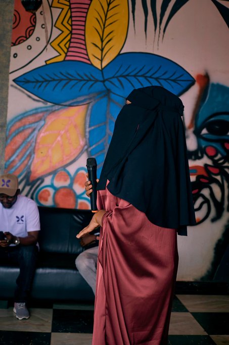 A woman in a black niqab and dusty rose dress speaks into a microphone while standing in front of vibrant graffiti-style murals, with a man in a white Hurley cap seated on a couch checking his phone nearby.

The image captures a moment of real-world connection at what appears to be a Web3 meetup or community event, embodying the post's theme of how online friendships in the crypto space translate into meaningful in-person gatherings beyond just digital tokens and trading.