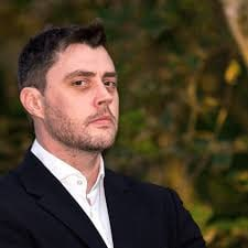 The image features a man wearing a suit, standing against a blurred background of trees. The man appears to be looking off into the distance or deep thoughtfully. The photo has been edited with a filter that gives it a vintage appearance, adding an artistic touch to the scene.