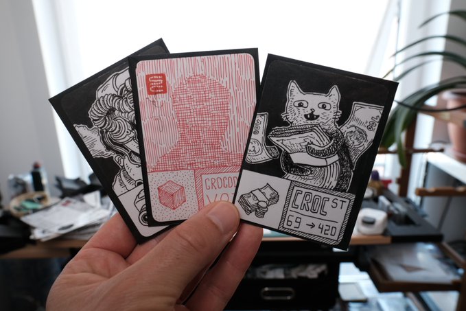 A hand holds up three illustrated cards/stickers including a cat character, a red crate labeled 'CROC', and an address 'CRO C ST 69 → 420'.