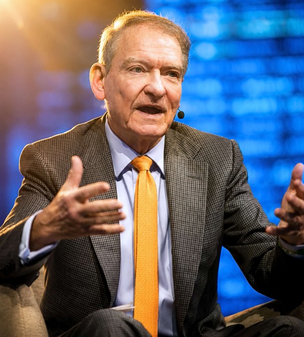 A photo of an older man in a checkered suit with an orange tie speaking while gesturing with his hands against a blue background.