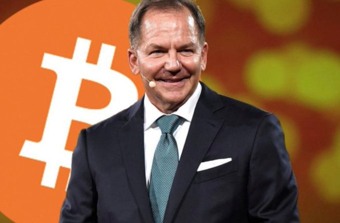 Paul Tudor Jones appears at a Bitcoin event, with a large Bitcoin symbol displayed behind him. The image complements news that his $106B Tudor Investment Corp. held $22.9M in BlackRock’s spot Bitcoin ETF (IBIT), up 18.9% quarter over quarter.