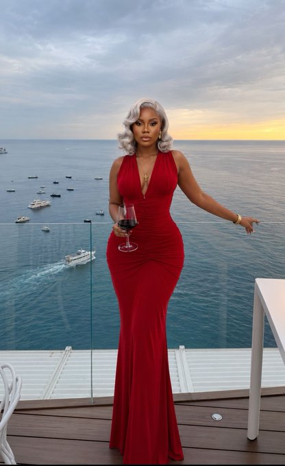 A woman with wavy platinum blonde hair wearing a fitted red halter gown holds a glass of red wine while standing on a balcony overlooking the ocean at sunset.