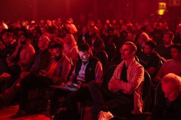 The image shows a large group of people sitting down, possibly attending an event or gathering. Some individuals are using their cell phones while others have laptops open before them. The room appears to be dimly lit with red lights, creating a cozy atmosphere for the attendees. A man in the foreground is wearing a vest and has his arms crossed over his chest. The post accompanying this image reads: "Day One at BTC Prague. And we're just getting started." This suggests that the event might be related to cryptocurrency or blockchain technology, possibly an ICO (Initial Coin Offering) or a similar gathering of crypto enthusiasts.