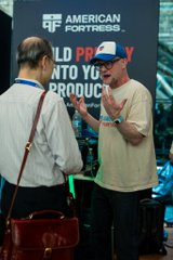 Two men in conversation at the American Portfolios booth, one wearing a beige shirt and cap, the other with a lanyard and shoulder bag.