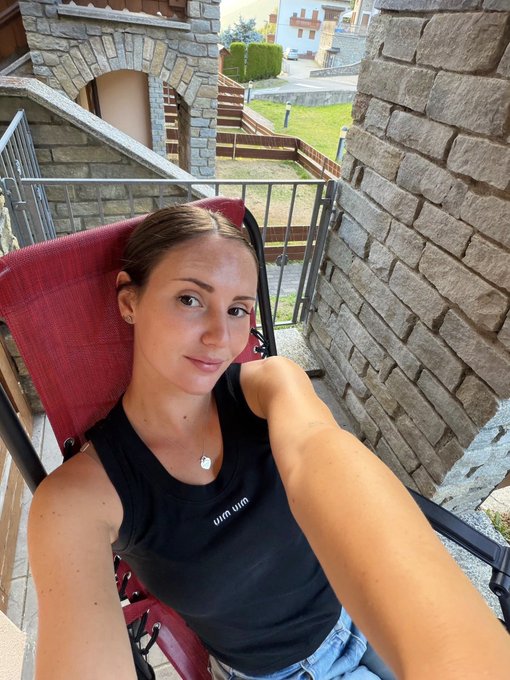 A selfie of a young woman with blonde hair sitting in a red folding chair on a balcony, wearing a black tank top with the mirrored text 'uni wun'.