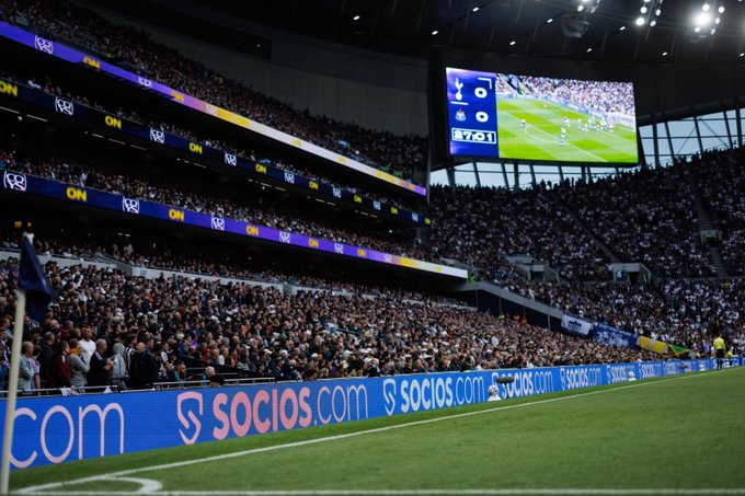A packed football stadium shows a match on the big screen with the score 0-0 between Spurs and Newcastle at 87:01, with 'socios.com' advertising boards visible along the pitch.