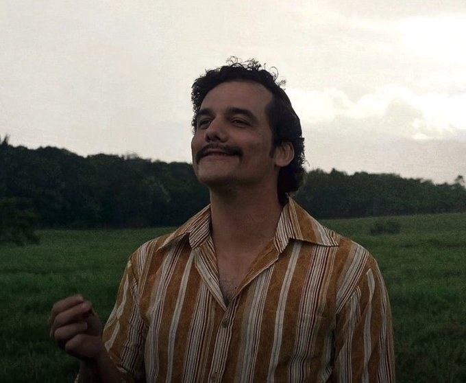 A man with dark curly hair and a mustache smiles softly while looking upward in a grassy field at dusk, wearing a striped orange and white button-up shirt.