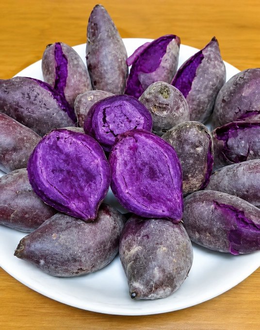 A white plate piled with whole and halved purple sweet potatoes, showing their vibrant violet flesh and dusty purple skin on a wooden surface.