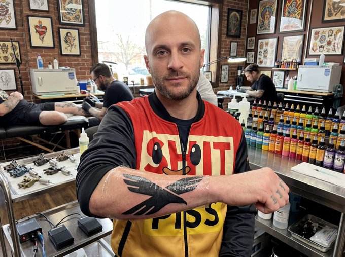 A bald man in a red and yellow shirt flexing his arm to display a freshly done bird tattoo in a tattoo parlor, with rows of colorful ink bottles and tattoo artwork visible in the background.
